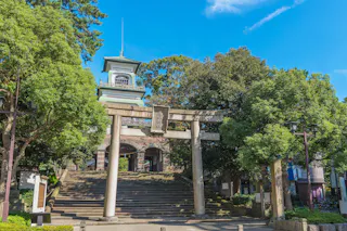 A traditional Japanese torii gate stands before stone steps leading up to a historic building, surrounded by lush green trees under a clear blue sky.