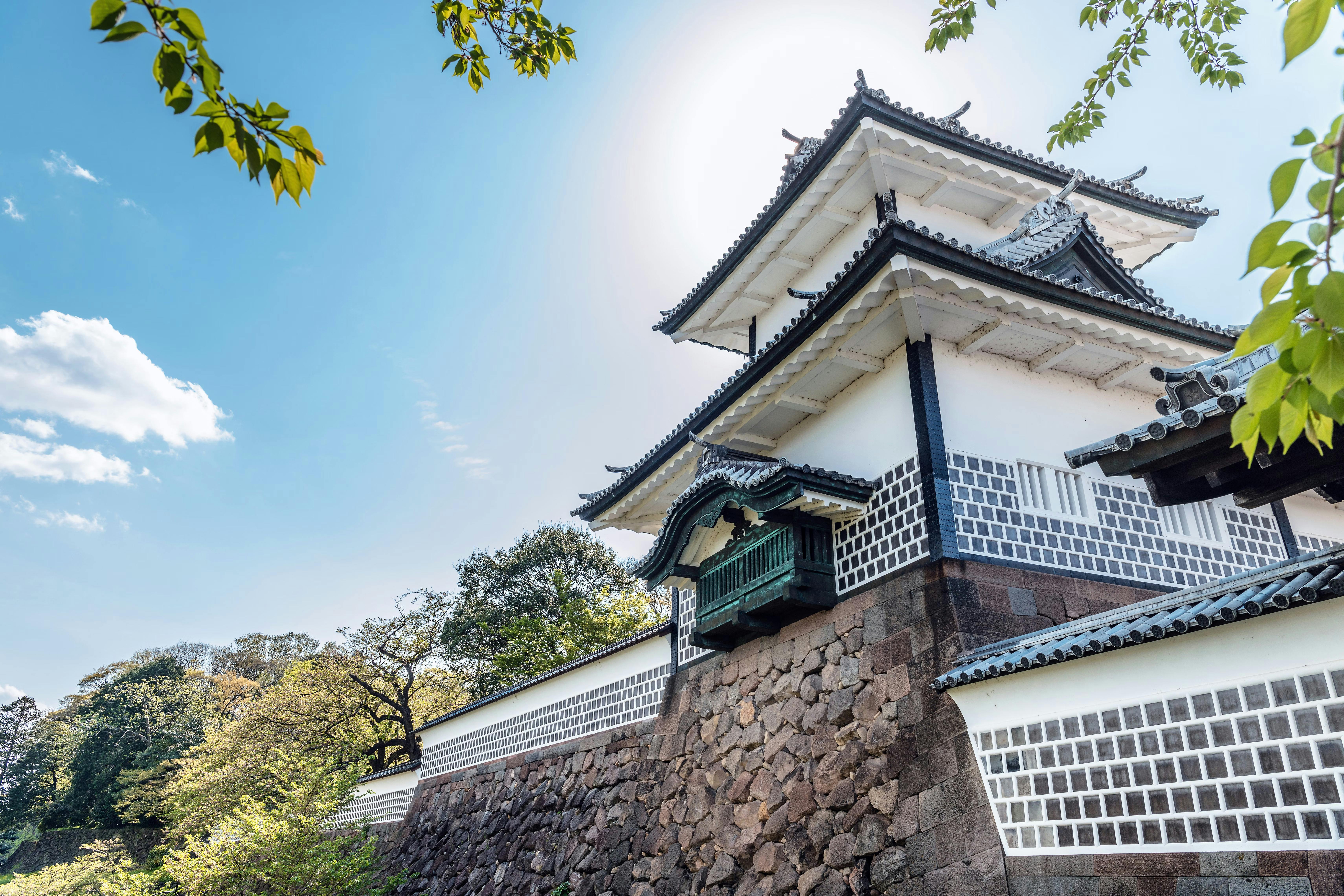 Traditional Japanese castle with white walls, dark tiled roofs, and a stone foundation, surrounded by trees under a bright blue sky with some clouds. Sunlight shines from behind the structure.