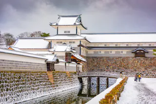 A traditional Japanese castle with white walls and roofs dusted with snow, surrounded by a stone wall and moat. Two people walk on a curved wooden bridge under a cloudy winter sky.