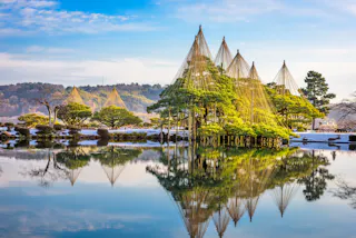 A tranquil Japanese garden with pine trees protected by conical rope structures, reflected in a calm pond under a blue sky, with distant hills and patches of snow visible in the background.