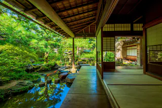 A traditional Japanese house with open sliding doors overlooks a serene garden with lush greenery, a pond, stepping stones, and stone lanterns under a wooden roof. Sunlight filters through the trees.
