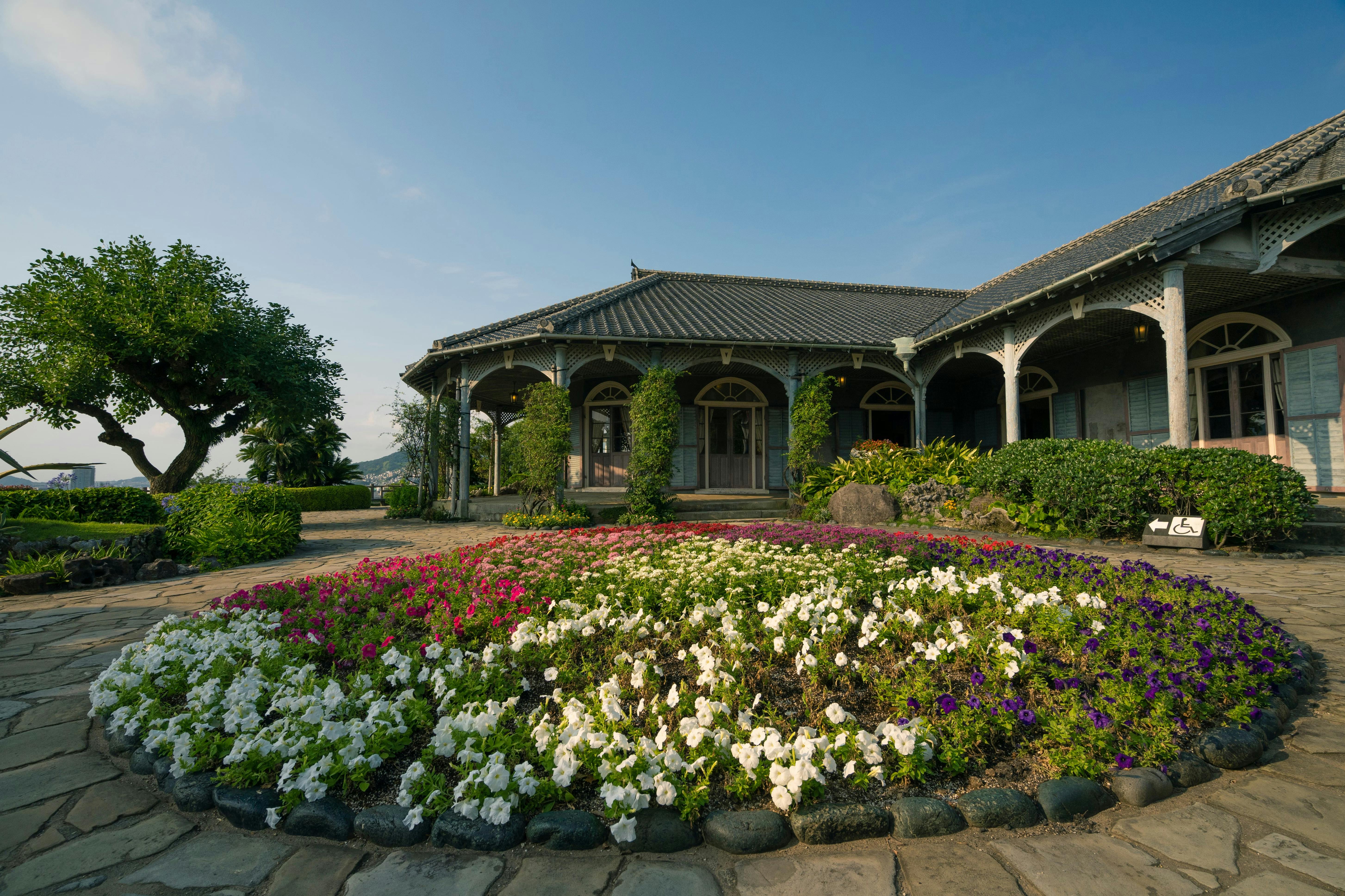 A historic wooden building with a wraparound porch sits behind a circular flower bed filled with colorful blossoms, surrounded by stone pavement and greenery under a clear blue sky.