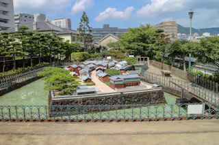 A detailed miniature model of a traditional Japanese village is displayed outdoors, surrounded by water and fencing, with modern buildings and trees in the background under a partly cloudy sky.