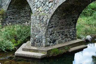 A close-up view of a stone arch bridge with a pillar standing in a shallow, clear stream, surrounded by green vegetation. The bridge is made of uneven, gray stones.