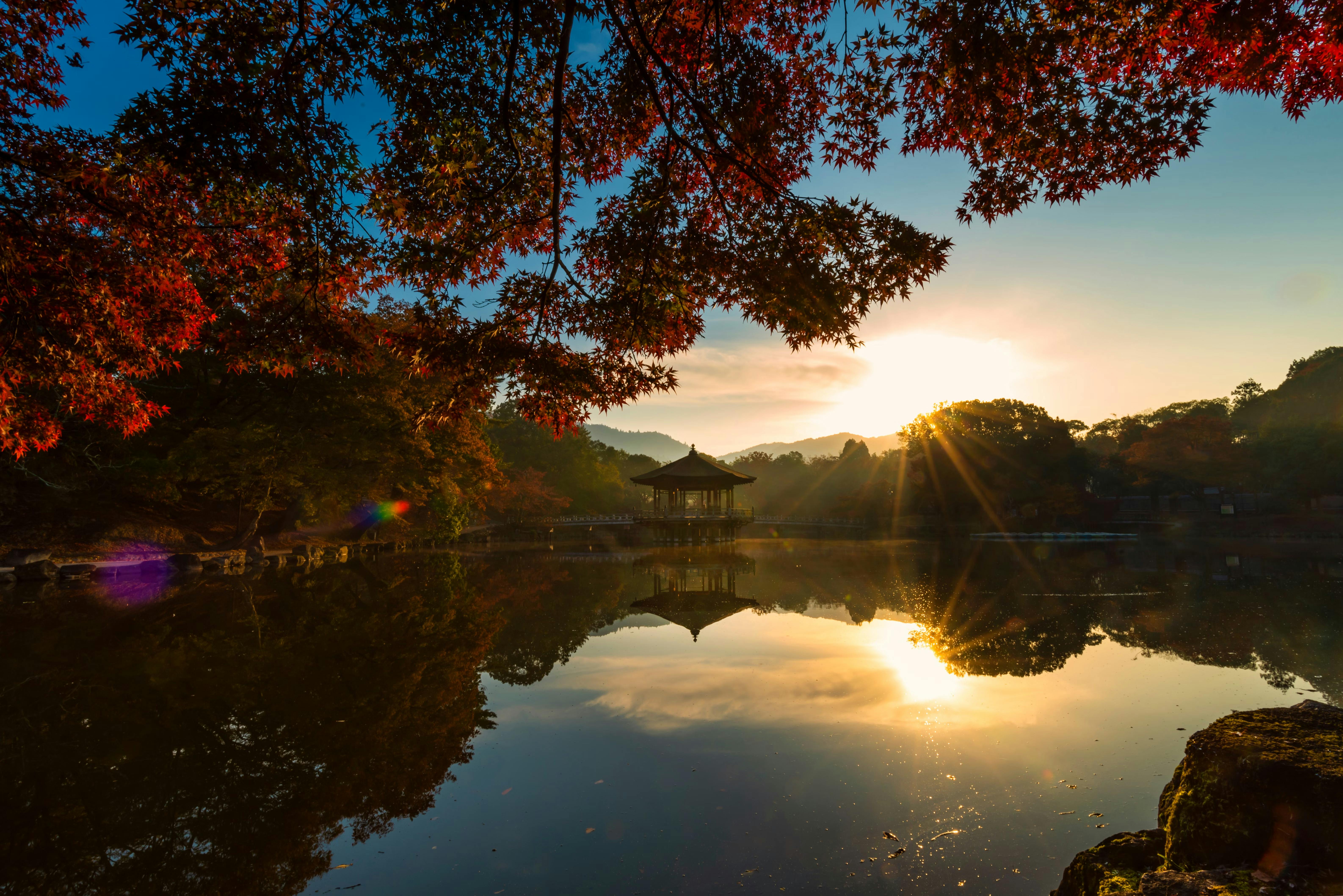 Sunrise over a calm lake with a traditional gazebo reflected in the water, framed by vibrant autumn leaves on tree branches. The soft sunlight creates a warm, serene atmosphere.