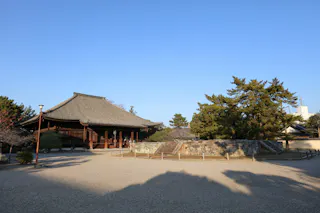 A traditional Japanese wooden temple with a sloped roof, surrounded by trees and a gravel courtyard under a clear blue sky.