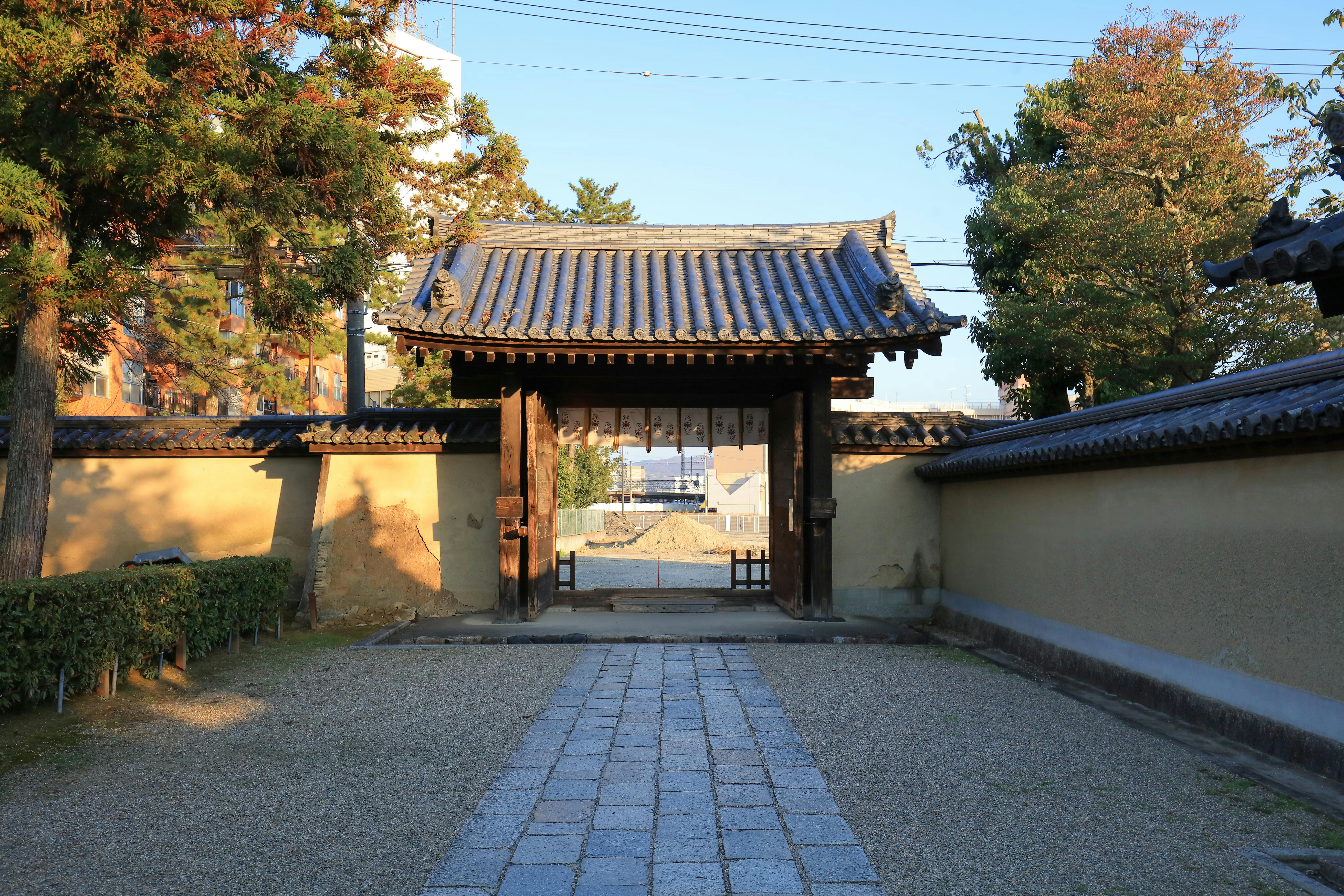 A traditional Japanese wooden gate with a tiled roof stands open, set in a tan plaster wall. Trees and a stone path frame the entrance, with blue sky and buildings visible in the background.