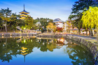 A tranquil pond reflects a traditional Japanese pagoda, trees, and nearby buildings at dusk in Nara, Japan. Soft lights illuminate the peaceful scene, surrounded by greenery and autumn foliage.