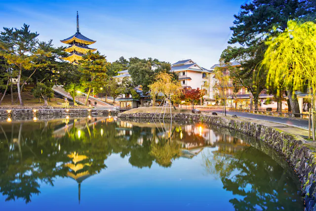 A tranquil pond reflects a traditional Japanese pagoda, trees, and nearby buildings at dusk in Nara, Japan. Soft lights illuminate the peaceful scene, surrounded by greenery and autumn foliage.