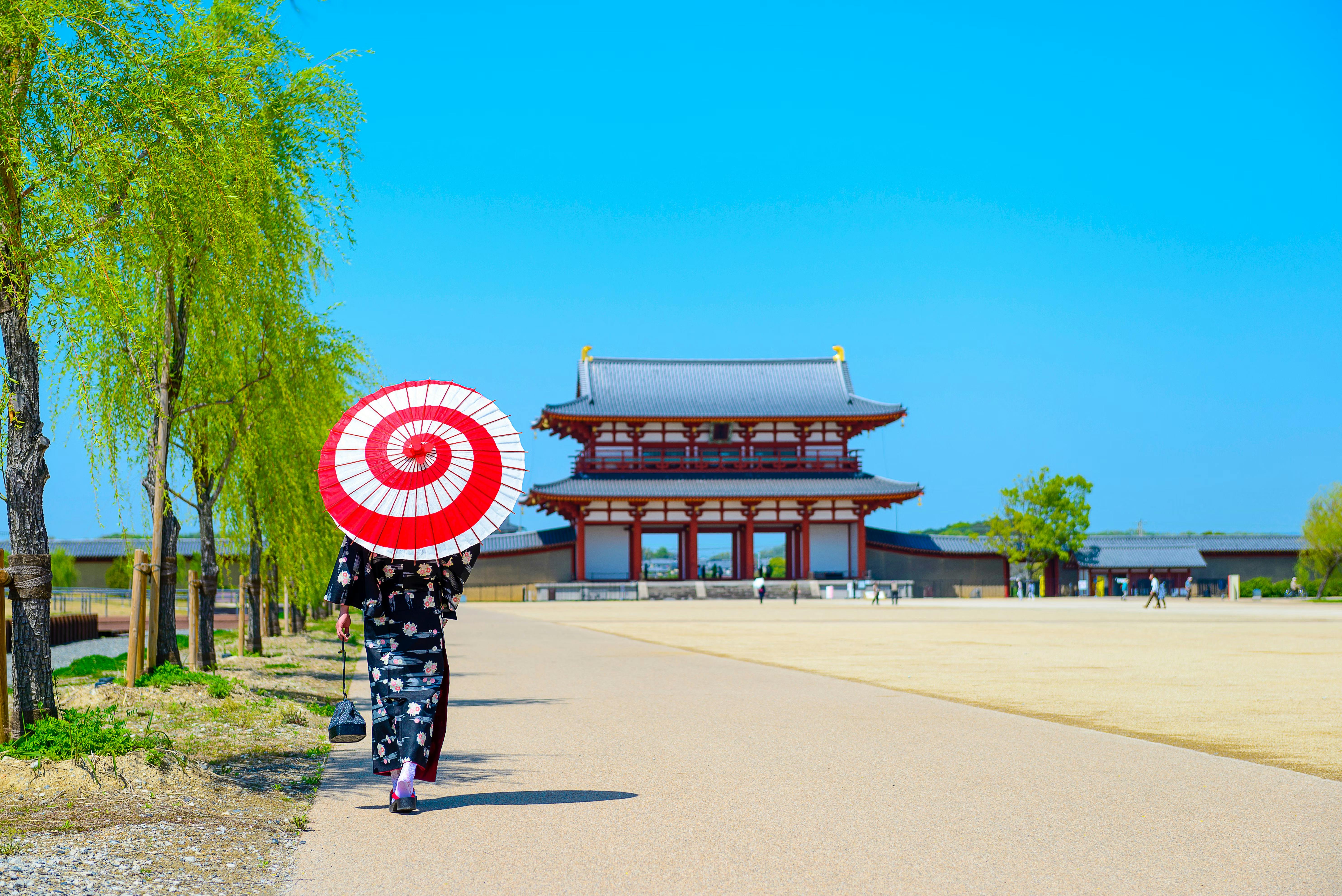 A person in a floral kimono walks on a wide path holding a red and white parasol, with a traditional Japanese temple building and blue sky in the background.