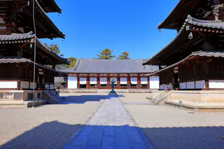 A traditional Japanese temple courtyard with wooden buildings on both sides and a main hall in the center, under a clear blue sky. Stone pathway leads to the main hall.