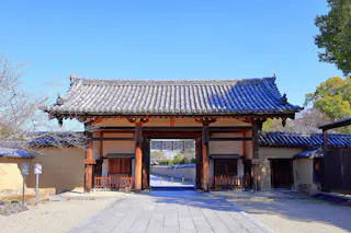 A traditional Japanese wooden gate with a tiled roof stands at the entrance to a historic site, flanked by stone paths and earthen walls, under a clear blue sky.