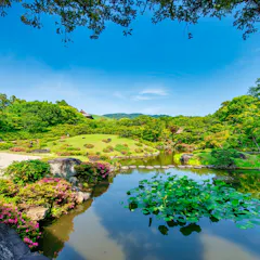 Isuien Garden A serene Japanese garden with a pond featuring water lilies, trimmed bushes, blooming flowers, rocks, and lush green trees under a bright blue sky with distant hills in the background.