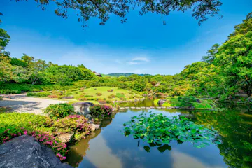 Isuien Garden A serene Japanese garden with a pond featuring water lilies, trimmed bushes, blooming flowers, rocks, and lush green trees under a bright blue sky with distant hills in the background.