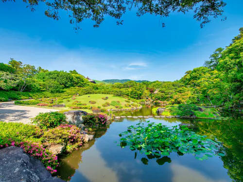 Isuien Garden A serene Japanese garden with a pond featuring water lilies, trimmed bushes, blooming flowers, rocks, and lush green trees under a bright blue sky with distant hills in the background.