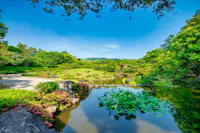 A serene Japanese garden with a pond featuring water lilies, trimmed bushes, blooming flowers, rocks, and lush green trees under a bright blue sky with distant hills in the background.
