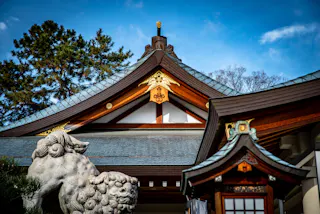 A traditional Japanese shrine with ornate wooden rooftops, gold accents, and a stone guardian statue in front, set against a blue sky and tall trees.
