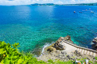 A rocky shoreline with clear turquoise water, a stone staircase leading down to the sea, lush green plants in the foreground, and several boats floating in the distance under a bright blue sky.