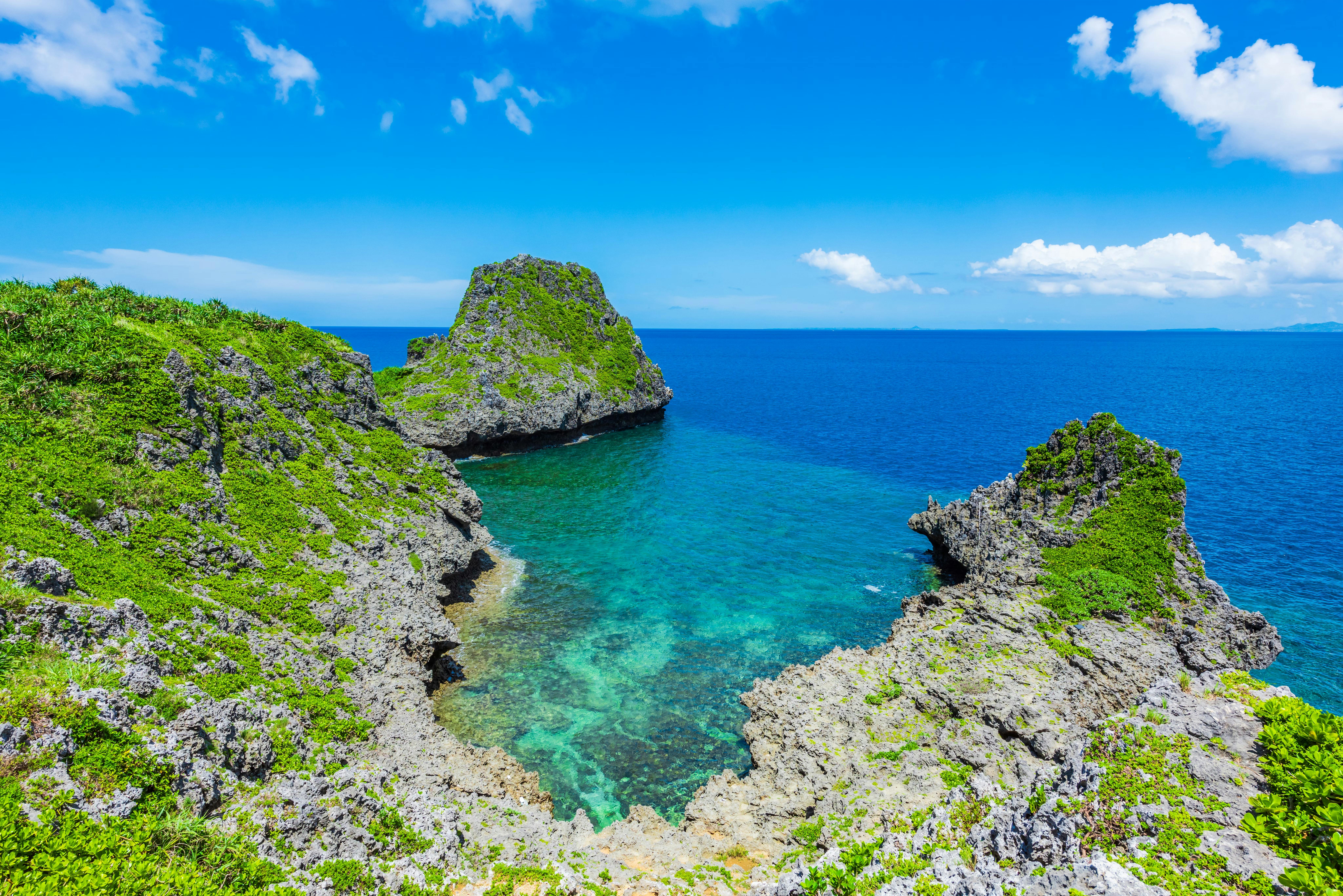 Clear blue ocean water surrounded by rugged, green rocky cliffs under a bright blue sky with scattered clouds. The vibrant landscape suggests a tropical coastal area.