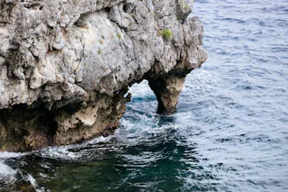 A natural rock formation with a small arch extends over clear, blue-green ocean water. Waves gently crash against the rocks, and some green vegetation is visible on the rocky surface.