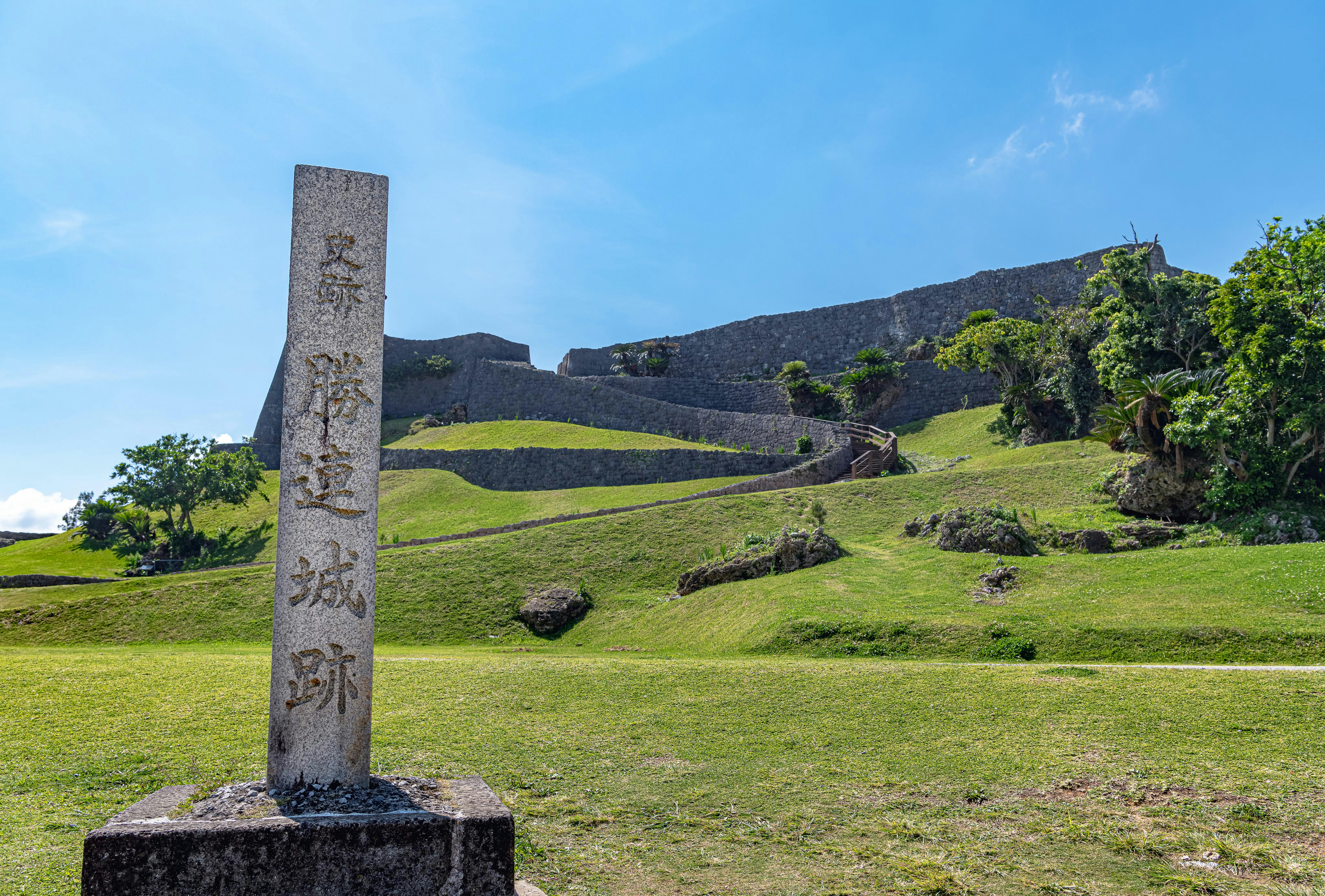 A stone marker with Japanese writing stands in front of an ancient stone wall and terraced grassy ruins under a blue sky, surrounded by greenery.