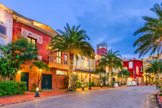 Colorful Mediterranean-style buildings with arched windows, palm trees, and warm lights line a cobblestone street at dusk under a blue sky.