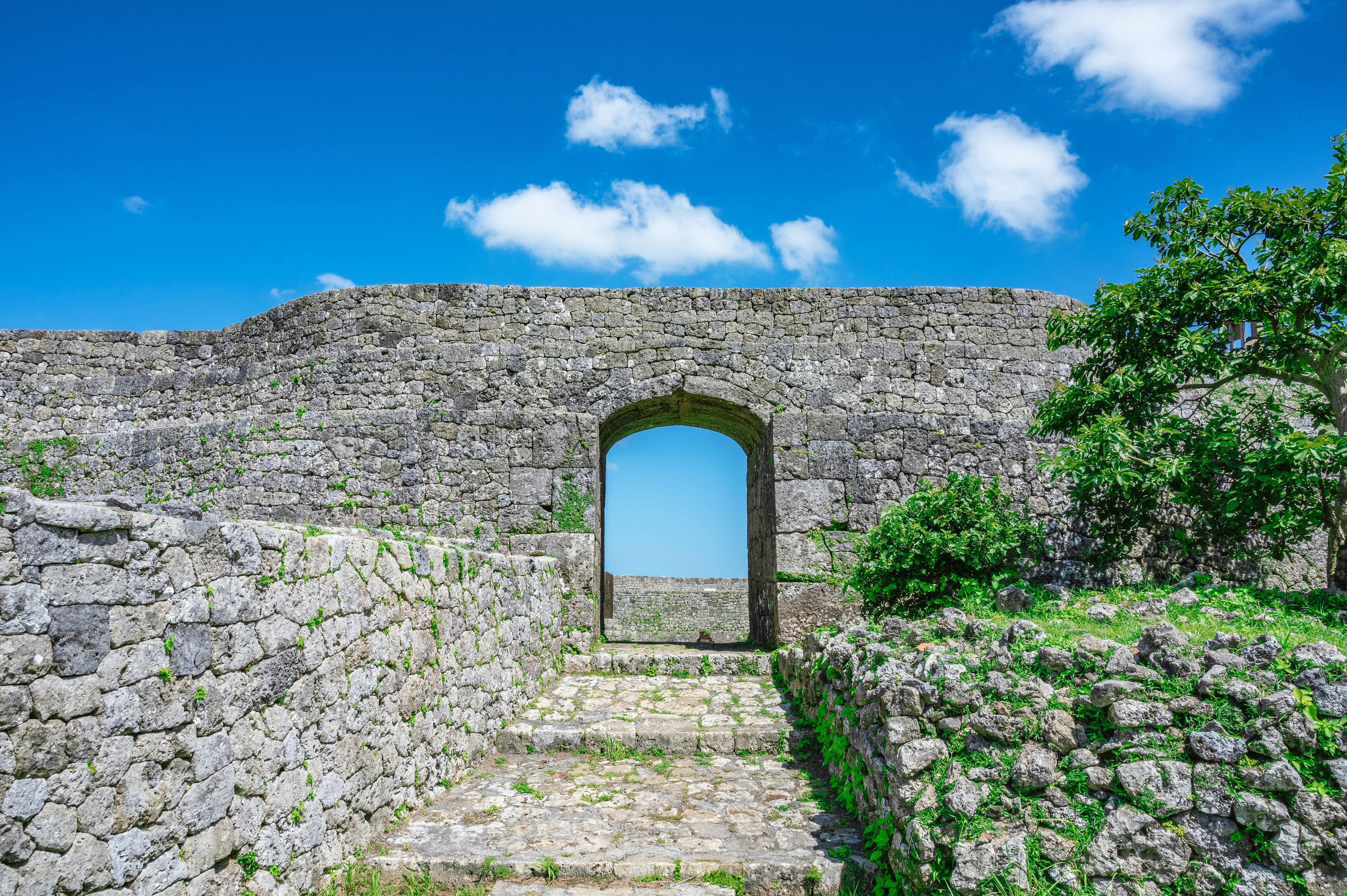 A stone archway in an old stone wall leads to a bright blue sky with a few clouds, surrounded by green grass and leafy trees under sunlight.