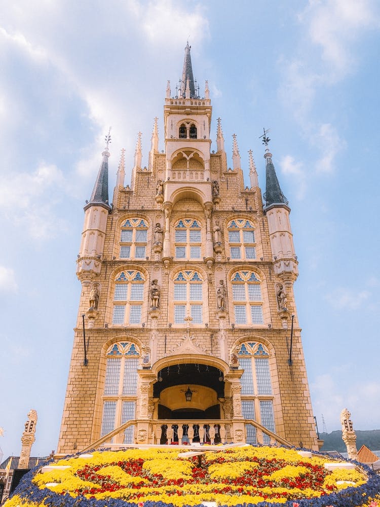 A grand, ornate building with tall spires and intricate stonework stands against a blue sky, with a colorful flower bed in the foreground and people gathered on the balcony.