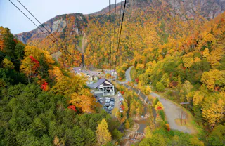 A cable car ascends over a colorful autumn forest with a parking lot, buildings, and winding roads below, surrounded by mountains covered in vibrant fall foliage.