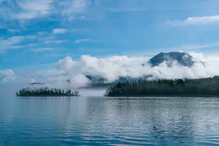A calm lake with reflections of the sky, low-lying mist over the water, a forested shoreline, and a mountain partially covered by clouds in the background under a blue sky.