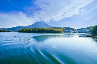 A serene lake with gentle ripples and clear blue water, surrounded by green trees and buildings on the shore. A mountain rises in the background under a partly cloudy sky.