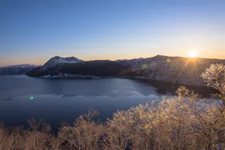 The sun rises over a mountain range beside a calm lake, with frosty trees in the foreground and a clear blue sky above.