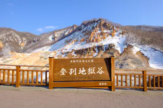 A wooden sign for Noboribetsu Jigokudani (Hell Valley) in Shikotsu-Toya National Park stands in front of a rocky, snow-dusted volcanic landscape with mountains in the background.
