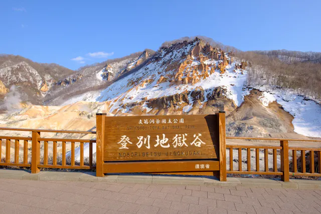 A wooden sign for Noboribetsu Jigokudani (Hell Valley) in Shikotsu-Toya National Park stands in front of a rocky, snow-dusted volcanic landscape with mountains in the background.