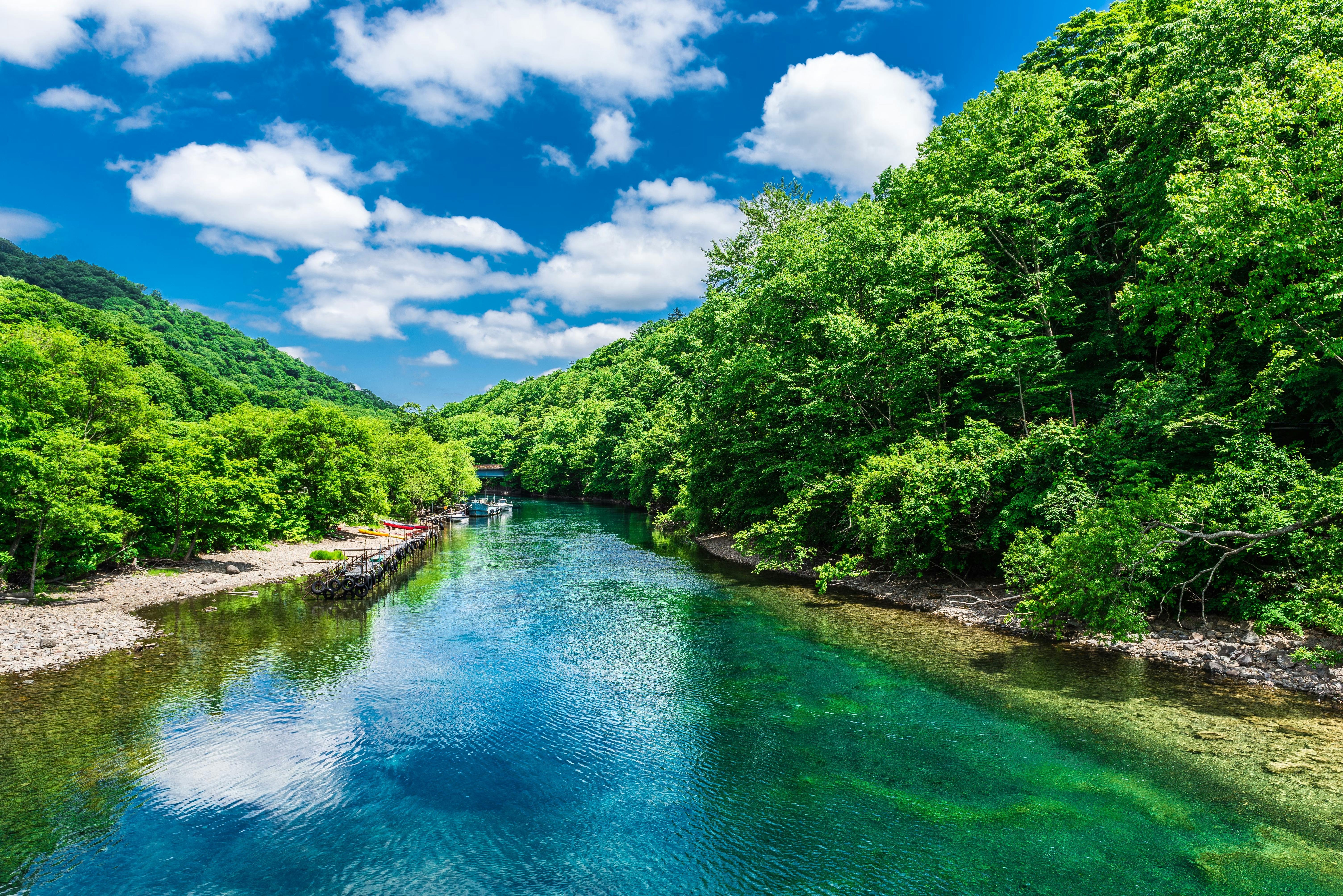 A clear river flows between lush green trees under a bright blue sky with scattered white clouds. The riverbanks are rocky with a few colorful boats docked on the left side.