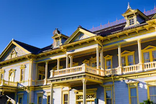 A large, ornate Victorian-style house with pale blue walls and elaborate yellow trim, featuring multiple windows, balconies, and decorative railings, set against a clear blue sky.