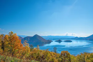 A scenic view of a blue lake surrounded by small forested islands and autumn trees with orange and yellow leaves, set against distant mountains and a clear, bright blue sky.