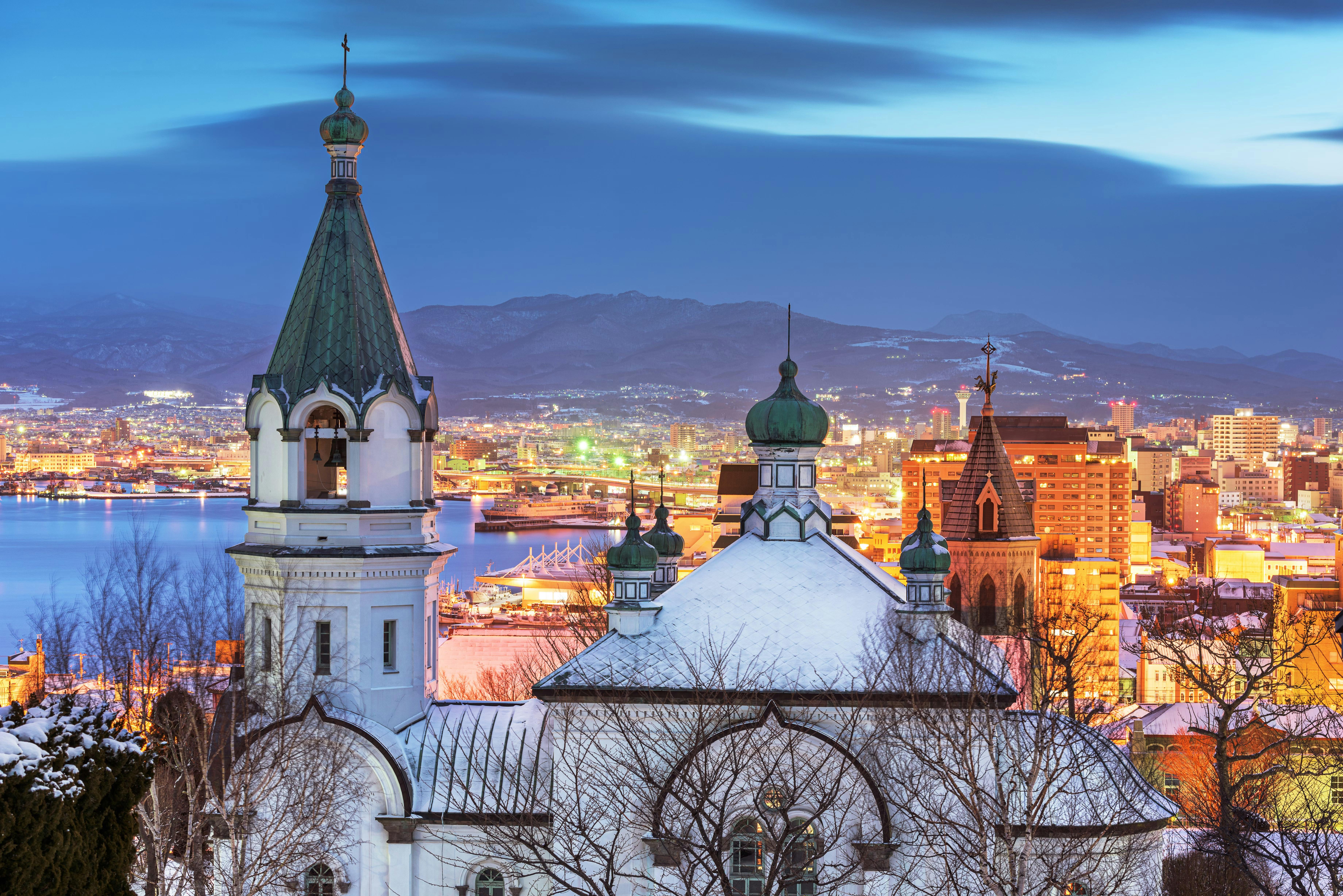 Snow-covered church towers in the foreground with a cityscape and illuminated buildings behind, set against mountains and a river at dusk under a blue sky.