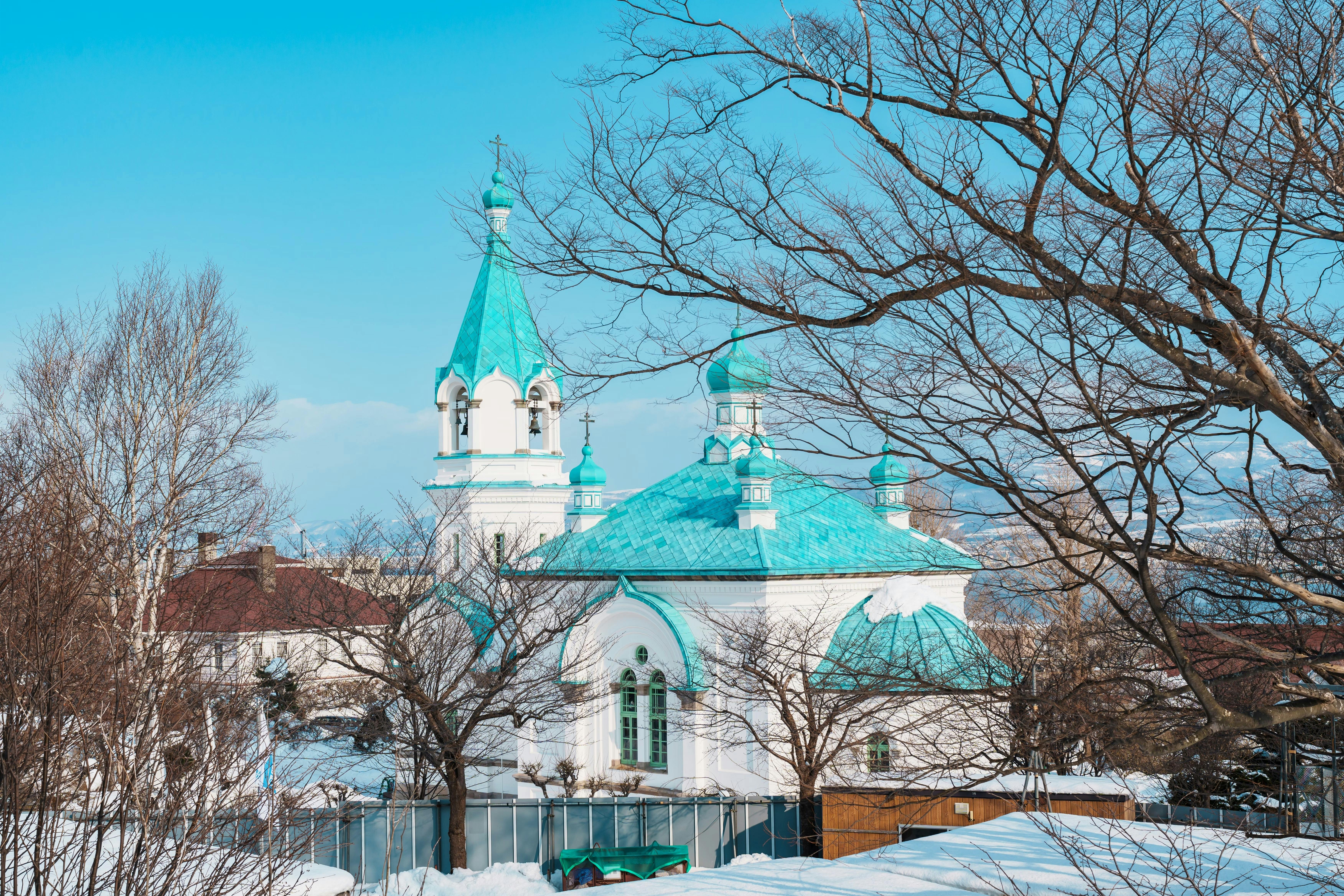 A white church with teal domes and spires stands among leafless trees in a snowy landscape under a clear blue sky.