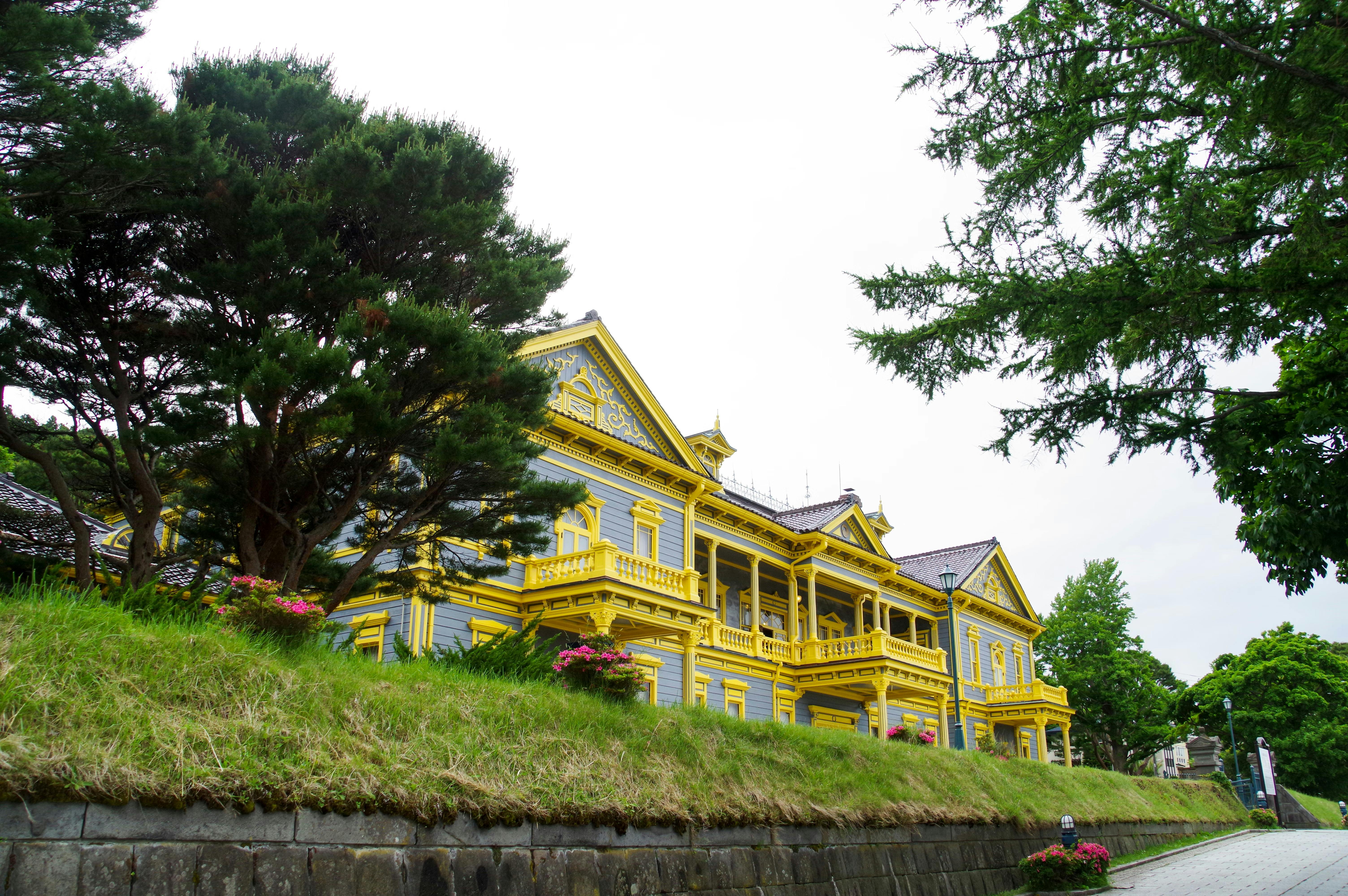 A large, ornate wooden building with bright yellow trim and balconies stands behind a grassy embankment and leafy trees under a cloudy sky.