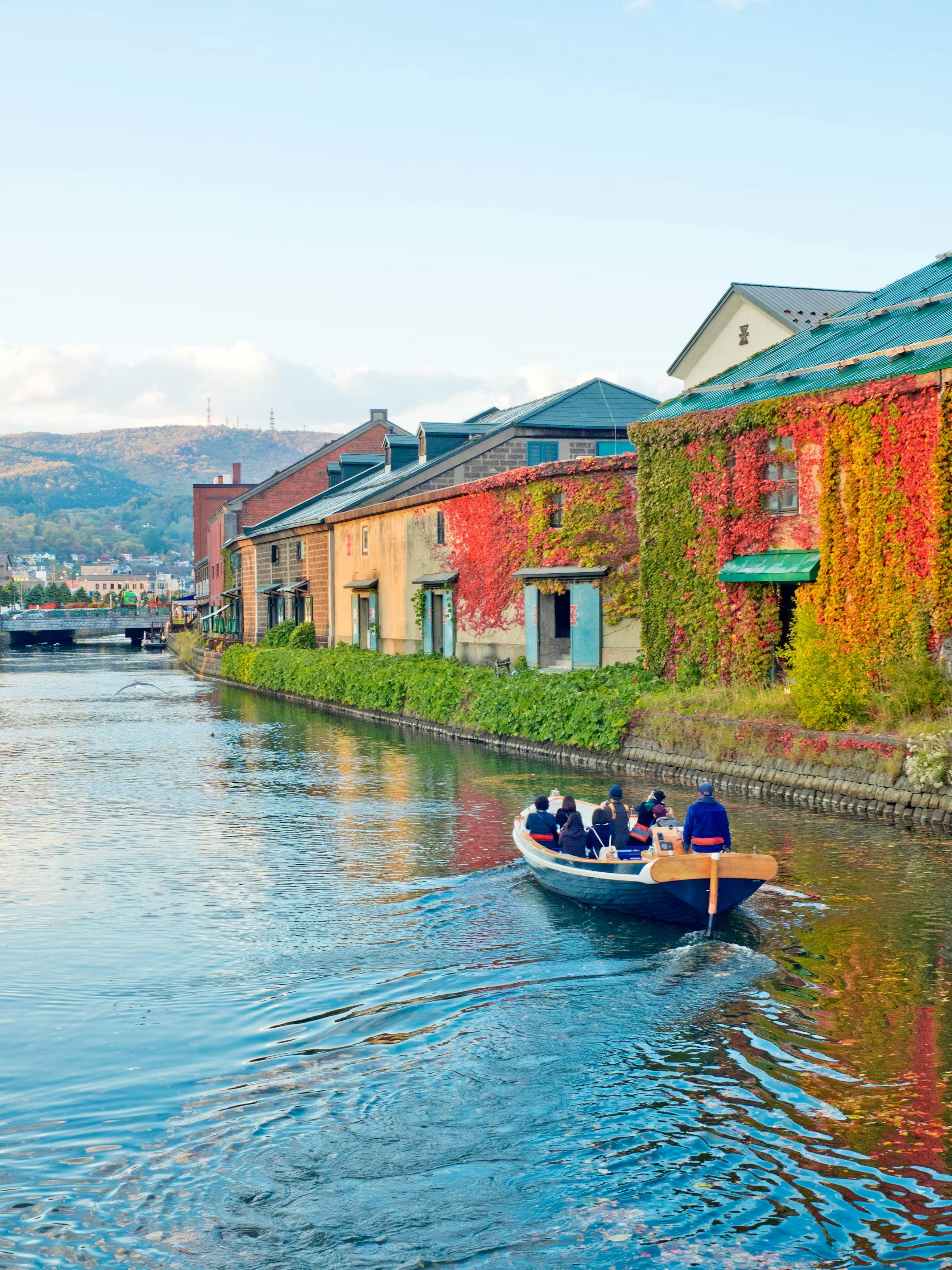A small boat with people rides along a calm canal lined with colorful ivy-covered buildings, with hills and a blue sky in the background.