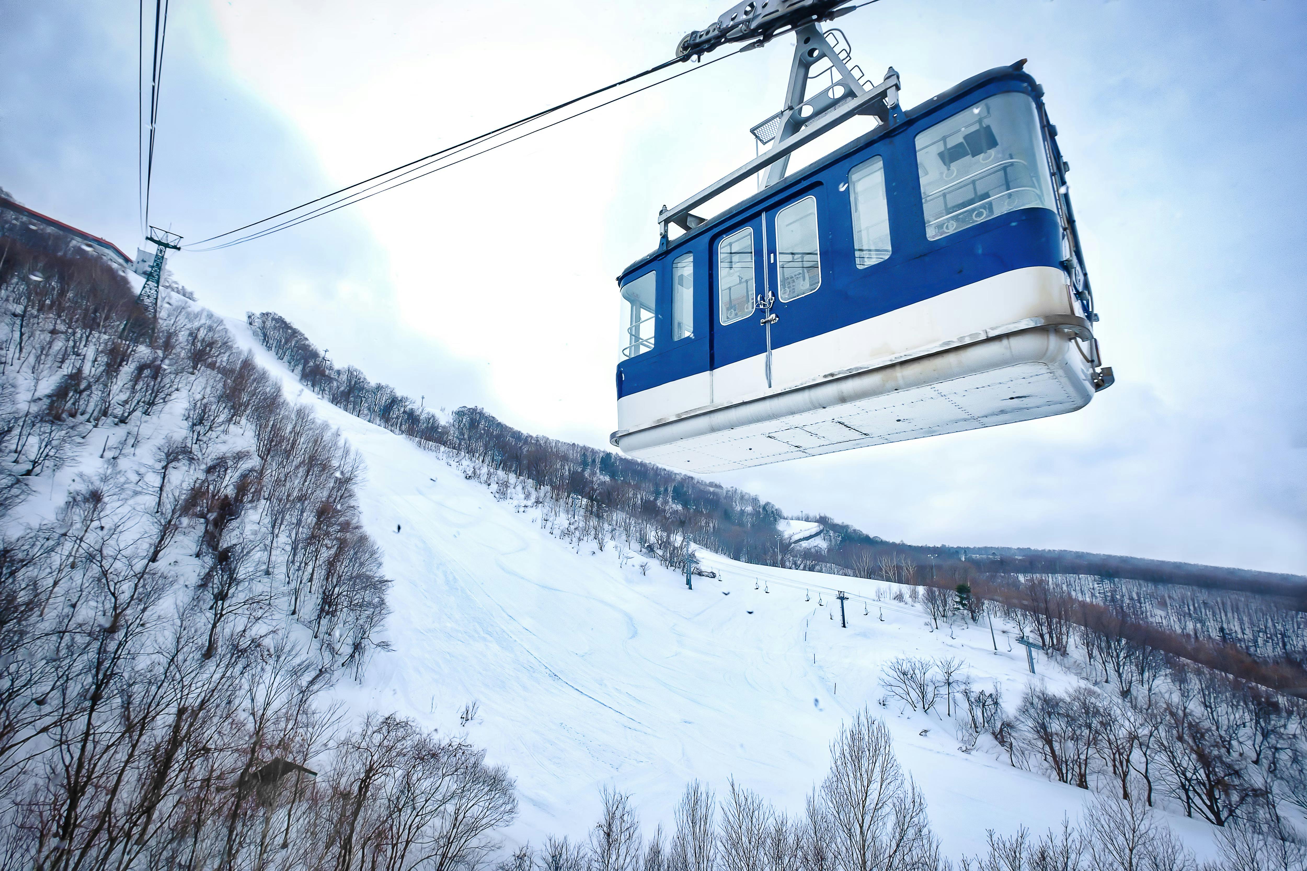A blue and white cable car travels above a snowy mountain landscape, surrounded by bare trees and ski slopes under a cloudy sky.