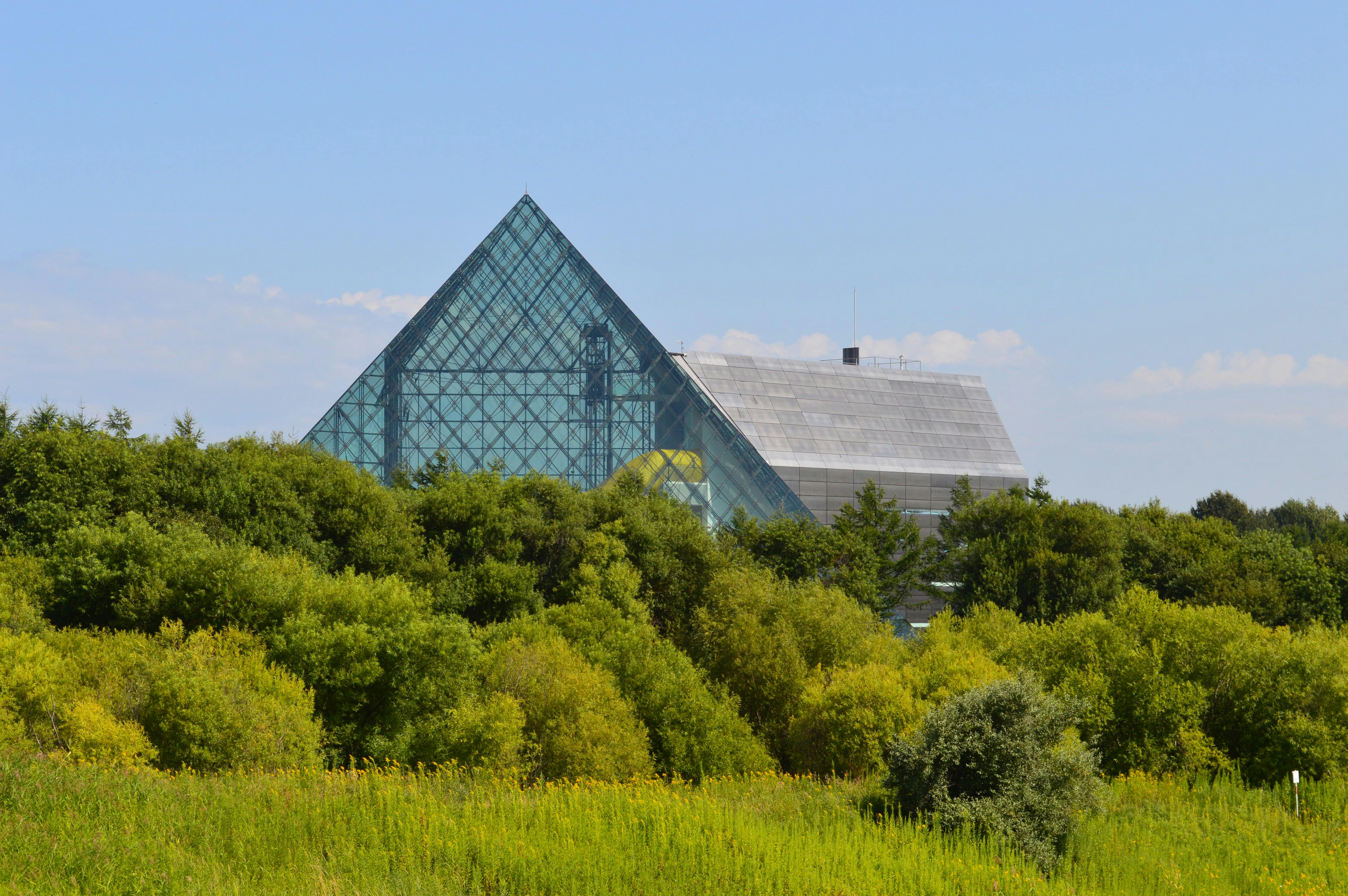 A large glass pyramid structure rises above green trees and shrubs on a sunny day, with a partly cloudy sky in the background.