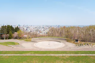 A circular stone amphitheater sits in a grassy park on a hillside, surrounded by trees, with a cityscape and tall buildings visible in the background under a clear blue sky.