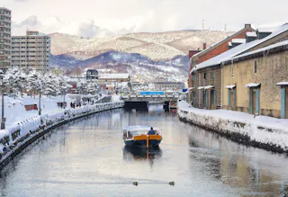 A boat moves along a snow-lined canal flanked by old warehouses, with people walking on the snow-covered path and distant mountains in the background under a cloudy sky.