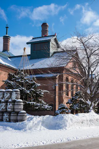 A red-brick building stands behind snow-covered trees and stacked sake barrels, with blue sky and clouds above. Snow blankets the ground, creating a bright winter scene.