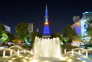 A brightly lit fountain at night with the illuminated Sapporo TV Tower in blue lights in the background, displaying the number 67. Trees and city buildings surround the scene.