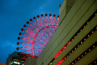 A brightly lit red Ferris wheel rises behind a modern building with horizontal rows of glowing lights, set against a deep blue evening sky.
