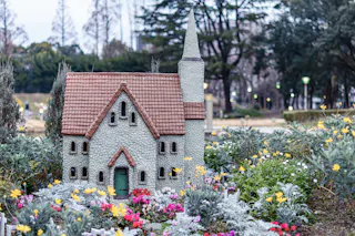 A small, detailed model of a stone church with a tall steeple sits among colorful flowers and greenery in an outdoor garden setting, with trees visible in the background.