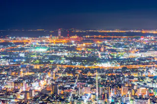 A vibrant cityscape at night, filled with brightly illuminated buildings and streets, stretches into the distance under a dark blue sky with a glowing horizon and scattered lights.